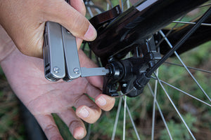 Close-up of a person using a Roxon multitool with a tire lever attachment to perform maintenance on a bicycle wheel outdoors.