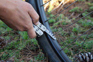 Close-up of a person using a Roxon multitool with a tire lever attachment to perform maintenance on a bicycle wheel outdoors.