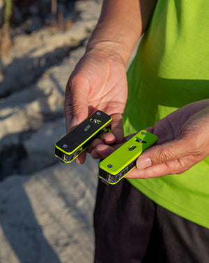 Two green and black devices held by a person on a sandy background
