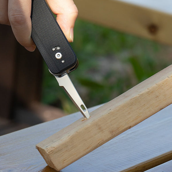 Hand holding a black folding knife with a blade inserted into a wooden plank outdoors.